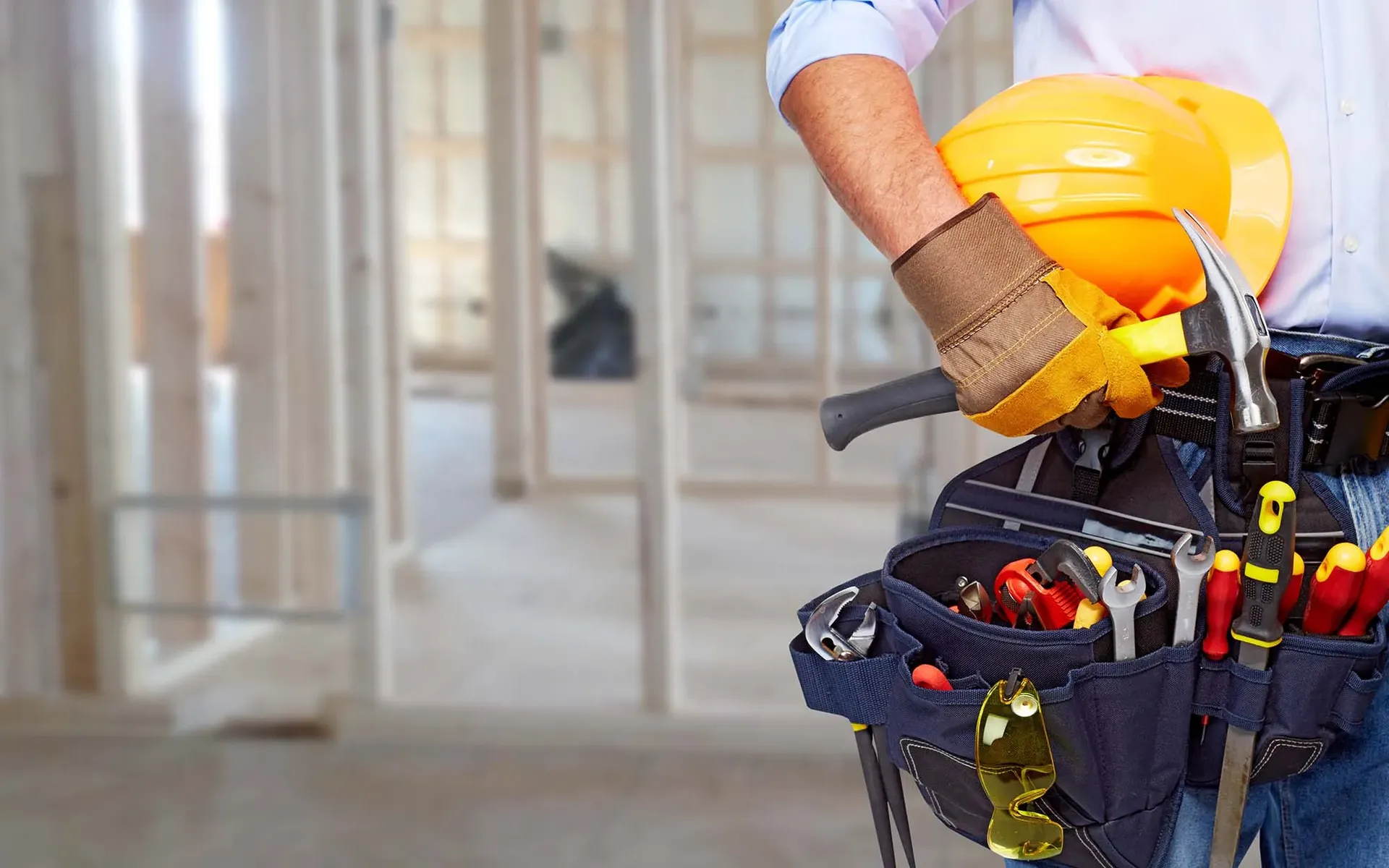 A worker wearing a toolbelt holds a hard hat at a construction site.