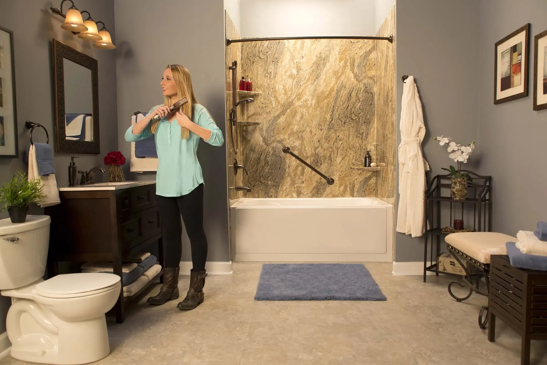 A person straightens their hair in a gray bathroom with a combination shower bathtub with brown marble-patterned walls, corner shelves, and black fixtures.