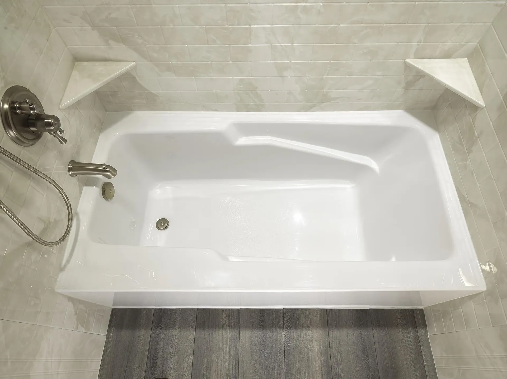 An overhead view of a white bathtub with off-white tiled walls and built-in corner shelves.