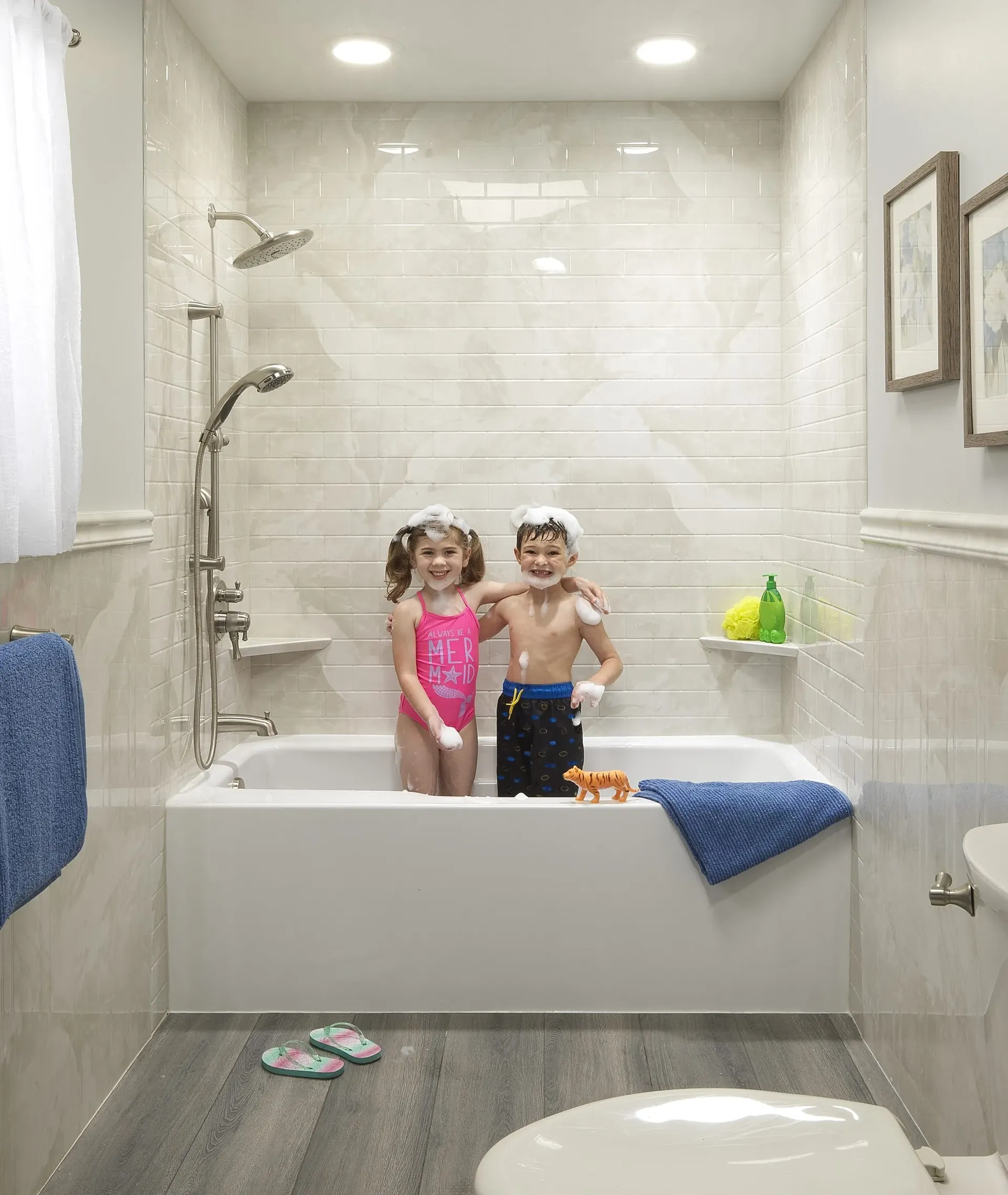 Kids in bathing suits covered in bubbles stand in a combination shower bathtub with off-white tiled walls.