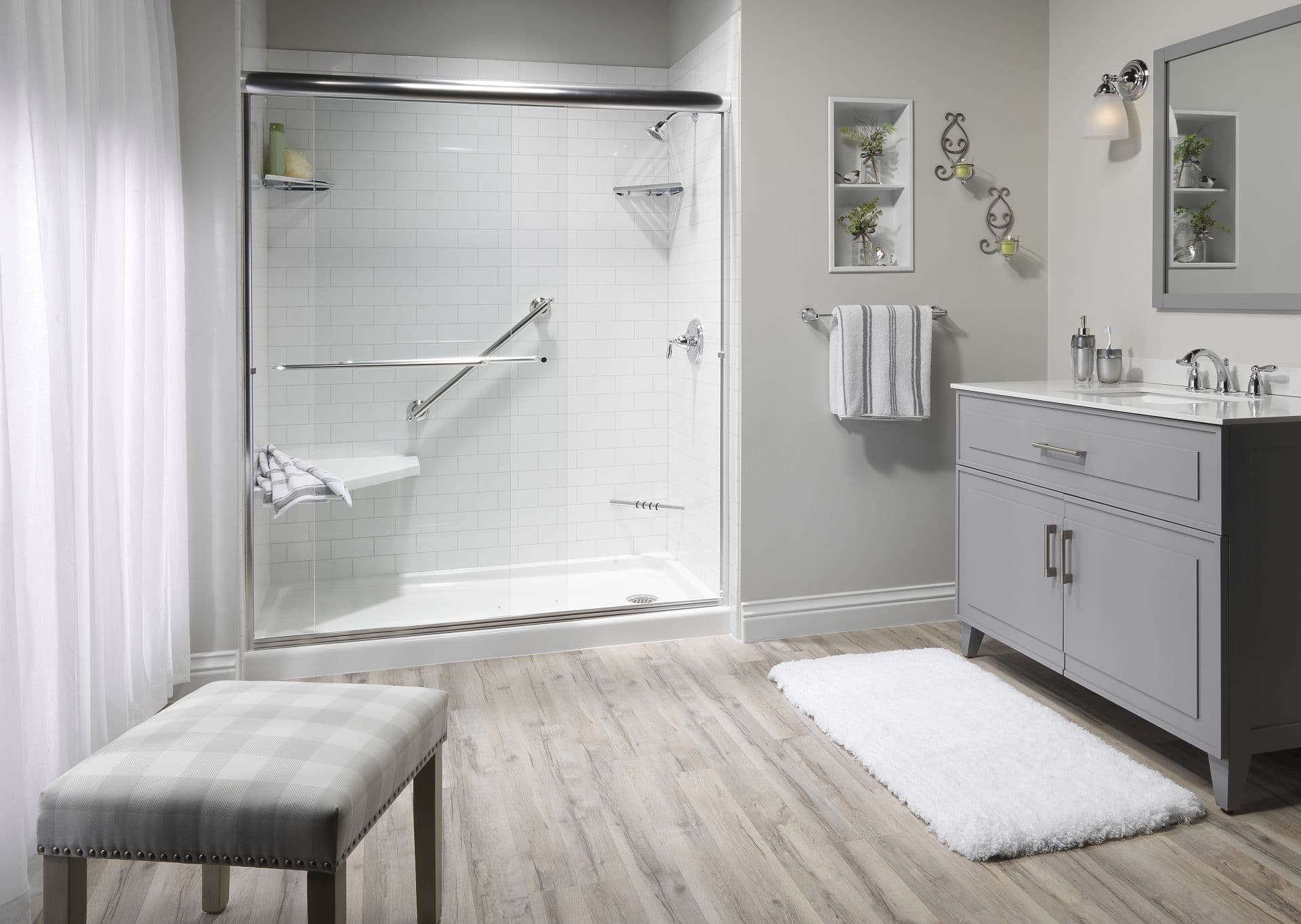 A light gray bathroom with a gray vanity, walk-in shower with sliding glass doors, diagonal grab bar, and built-in corner bench seating.