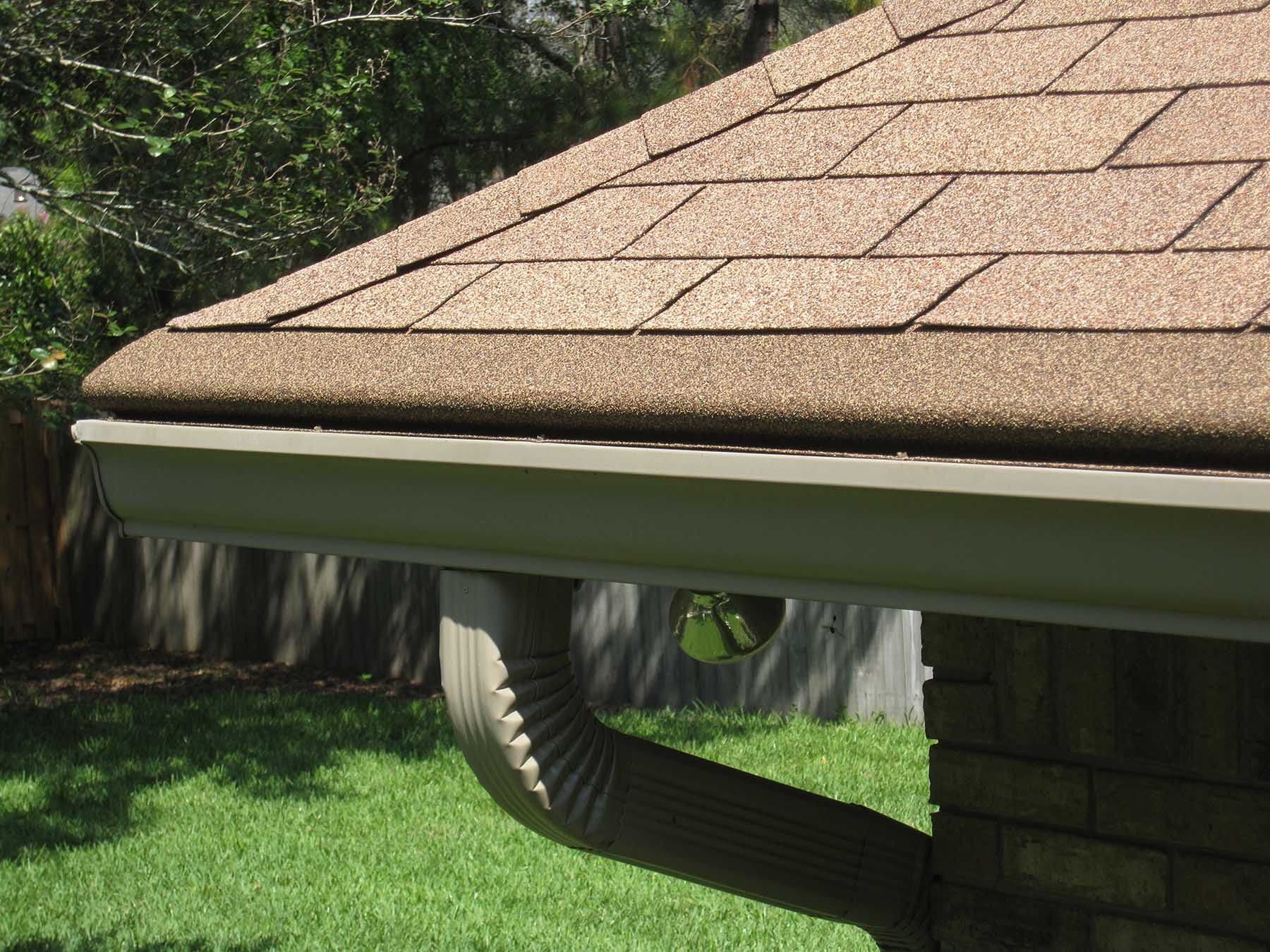 A close-up of the corner of a house with new gutters and light brown shingle roofing.