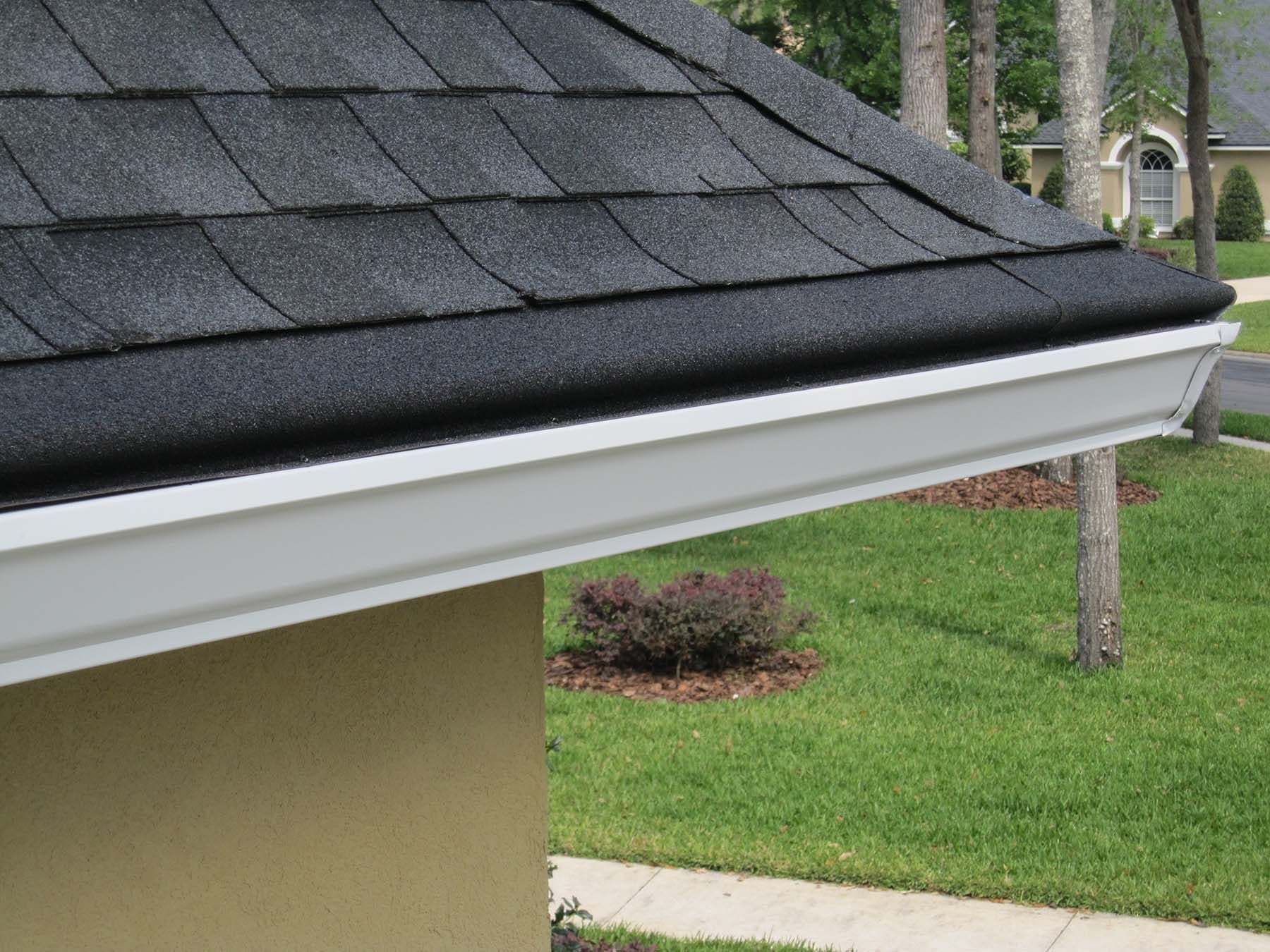 The corner of a tan house with black roof and new white gutters with cover.