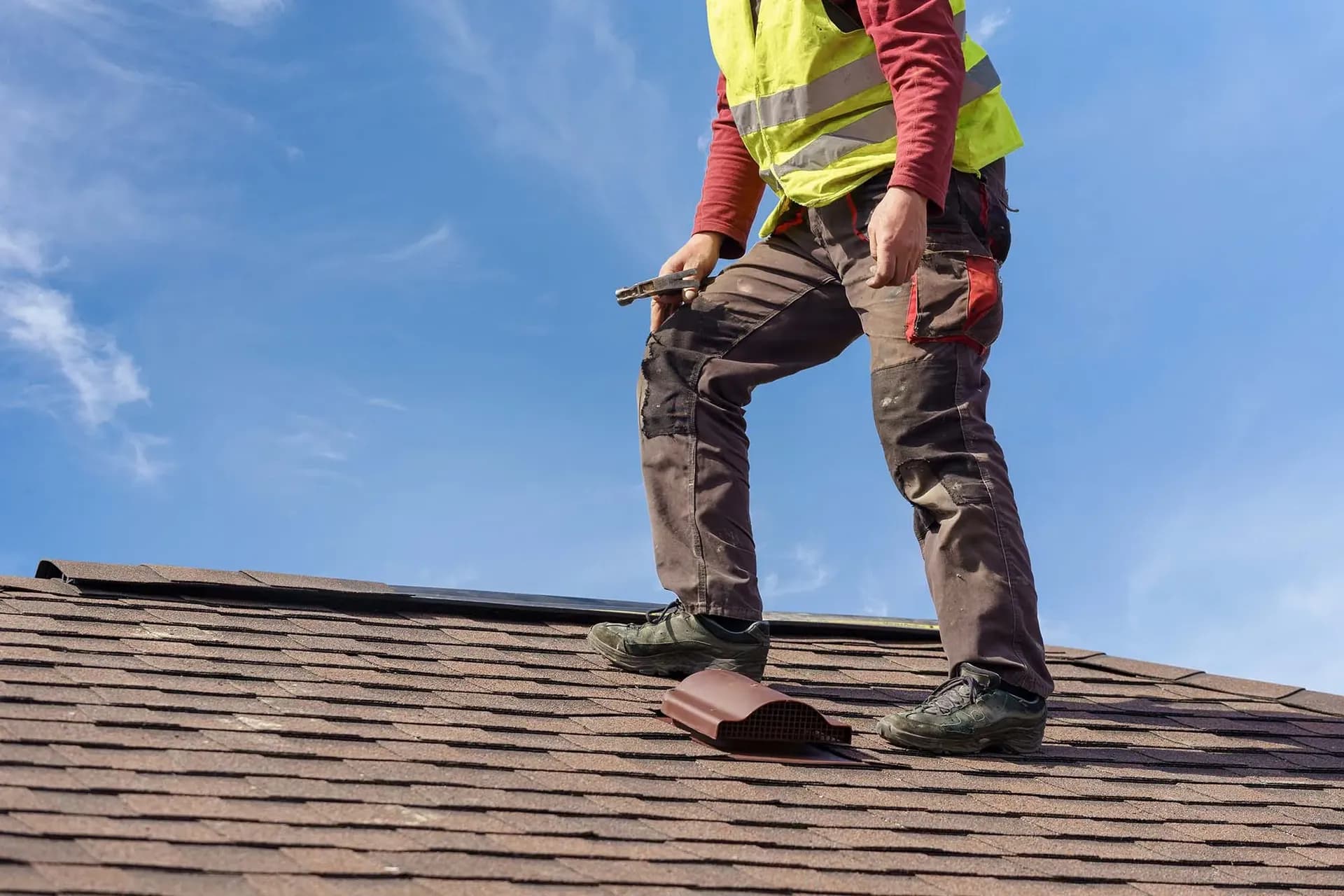 A worker in a safety vest stands on a roof holding a hammer.